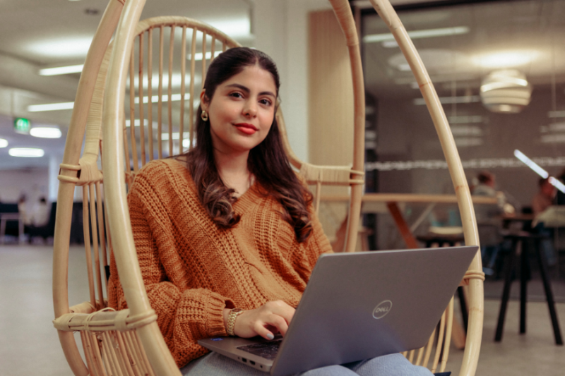 A students with a laptop sitting in a wicker chair at the university, smiling at the camera